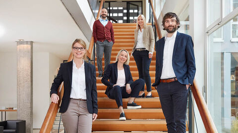 Fünf unserer Kolleginnen und Kollegen stehen beziehungsweise Sitzen auf einer Treppe.